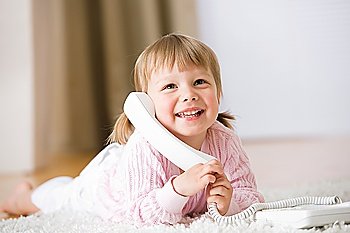 Smiling little girl lying down on carpet with phone calling holding telephone receiver at home