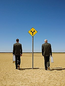 Two businessmen with briefcases walking past road sign in desert back view