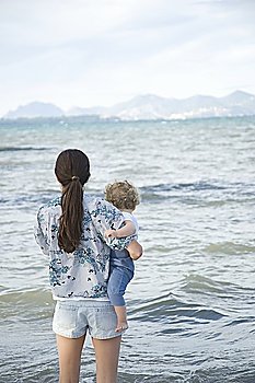 A lady holding a toddler standing in front of the sea with mountains in the far background