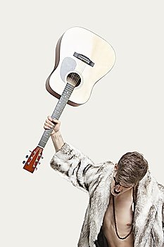Frustrated young man in fur coat about to throw his guitar against gray background