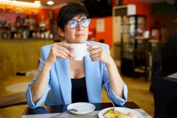 Thoughtful middle aged female in blue jacket and eyeglasses sitting at table with cup of hot beverage and looking away while resting in cafe Pensive 
