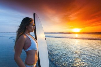 A beautiful young slim sporty woman in bikini with a surfboard is standing at ocean beach at sunset Woman with surfboard