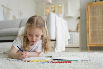 Talented little preschool child girl painting lying on warm floor in living room at home Caucasian kid girl drawing with colored pencils playing spen