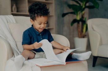 Happy mixed race little kid boy with cute curly hair reading story book while sitting in cozy armchair in light colored living room at home  child spe