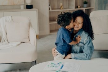 Cute mixed race boy little kid gently hugs his happy mother sitting on floor by table with colored pencils on it  surrounded by light colored furnitur