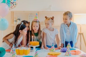 Small kids celebrate birthday party  blow candles on cake  gather at festive table  have good mood  enoy spending time together  make wish  wear party