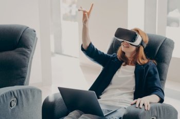 Young impressed redhead businesswoman using virtual reality glasses for business  female in VR headset pointing with finger in air  touching 3d object
