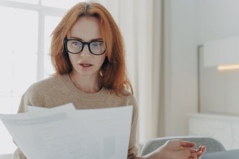 Serious redhead young woman focused in paper documents studies financial report wears spectacles casual jumper poses indoor against home interior Bus