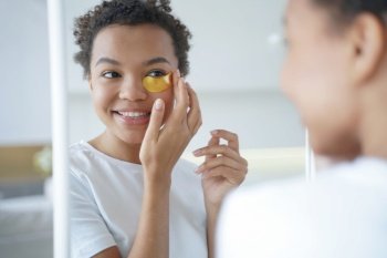Mirror reflection of young hispanic lady Happy afro girl is applying eye patches to her face Skin moisturizer with collagen serum Anti age patches