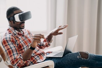 Smiling african american man in VR glasses sits in armchair with laptop  testing virtual reality goggles Modern black guy viewing purchase in cybersp