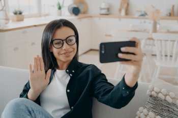 Smiling young female greeting friend  waving hand  having video conversation by smartphone sitting on couch at home Businesswoman wearing glasses cha