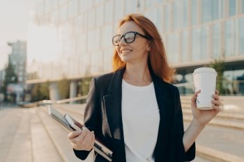 Overjoyed happy ginger woman in spectacles drinking coffee and holding laptop while walking outdoors on sunny day  business woman enjoying her way to 