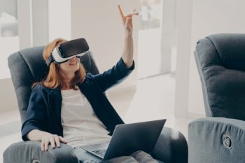 Impressed redhead businesswoman using VR glasses for remote work  interacting with 3D objects and pointing in the air while working on her laptop from