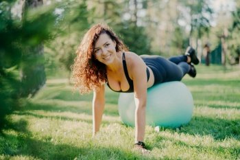 Happy sportswoman in active wear uses gymnastic ball for outdoor fitness  posing on green grass with a smile