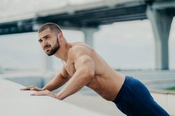 Bearded man does push-ups outdoors  warms up  poses near a bridge  has early morning workout