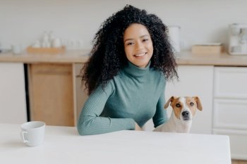 Curly-haired woman in turtleneck sits at kitchen table  enjoys tea  plays with Jack Russell terrier Joyful moment with beloved pet