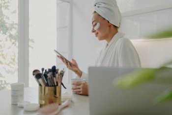 Morning routine Businesswoman in bathrobe  hair wrapped  under-eye patches  uses smartphone  prepares for work at home  kitchen background