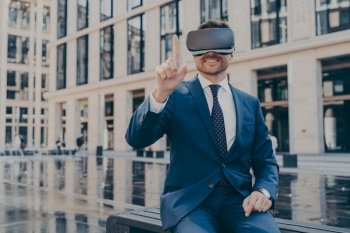 Happy businessman in blue suit testing VR goggles on bench  controlling with finger Excited man outdoors  office buildings blurred