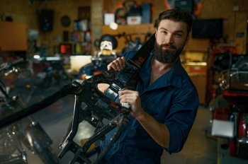 Portrait of young bearded repairman in coveralls carrying spare part of motorcycle frame on shoulder and looking at camera Motorcycle garage repair s