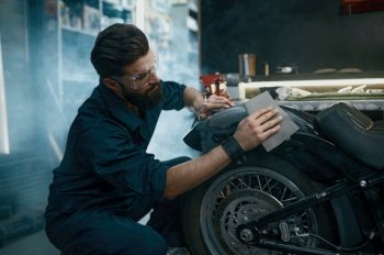 Young man worker wearing protective glasses sanding motorcycle from old paint while working at repair service Young man repair service worker sanding