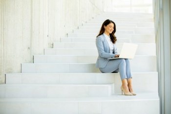 Pretty young business woman sitting on the stairs on office hallway and working on laptop