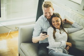 Smiling couple in casual outfit  sitting on the couch at the living room while watching something at mobile phone