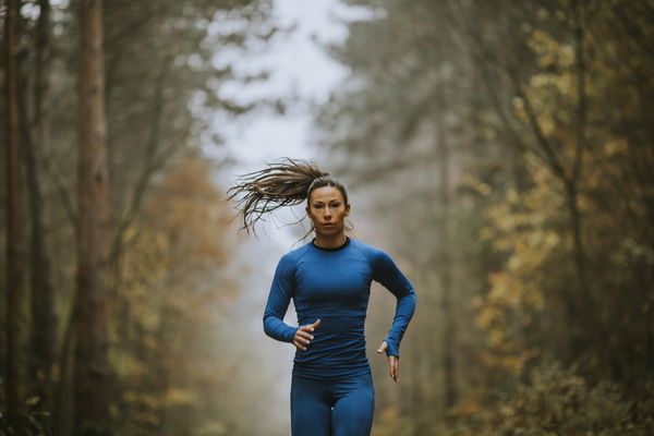 Young woman in blue track suit running toward camera on the forest trail at autumn