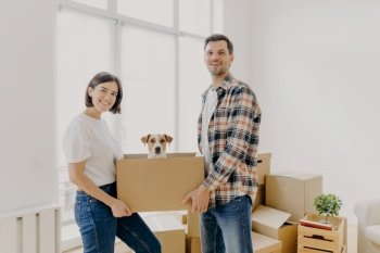 Happy family couple hold carton box with small puppy  stand indoor against big window  glad to become homeowners  unpack things in own apartment  look