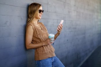 Glad female in stylish clothes and sunglasses  with cup of coffee smiling and browsing social media on cellphone while leaning on gray wall on city st