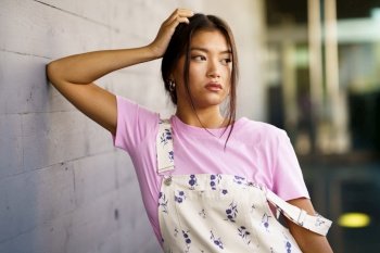 Young Asian woman in pink t shirt and stylish overalls touching head and looking away while leaning on gray wall on city street Stylish Asian female 
