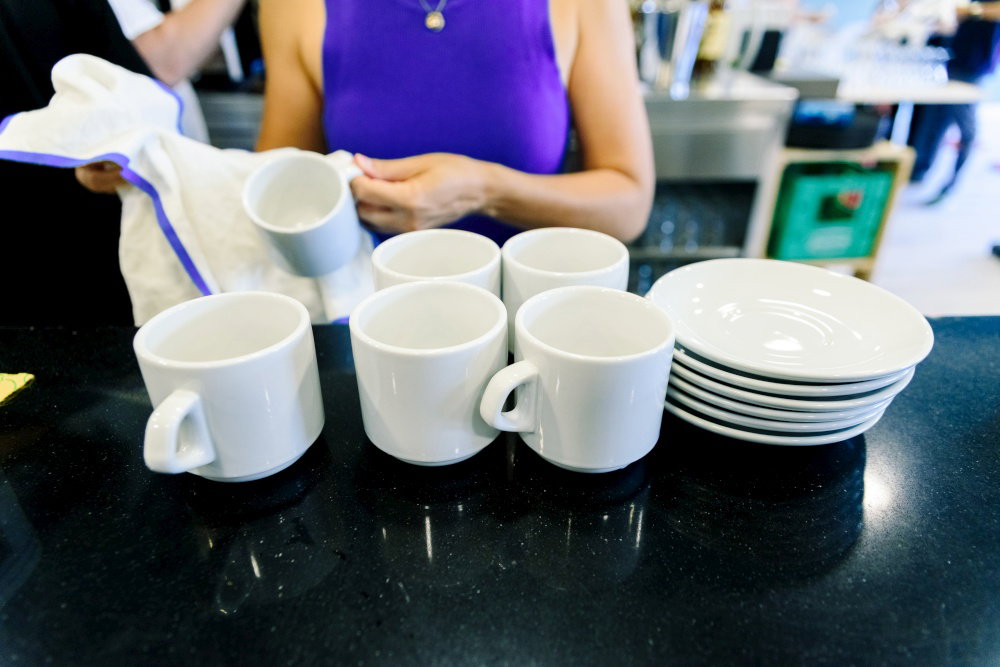 A waitress with dark hair wipes clean coffee cups and plates in a cafe She prepares for the next round of customers during the morning rush A waitr A waitress with dark hair wipes clean coffee cups and plates in a cafe She prepares for the next round of customers during the morning rush A waitr