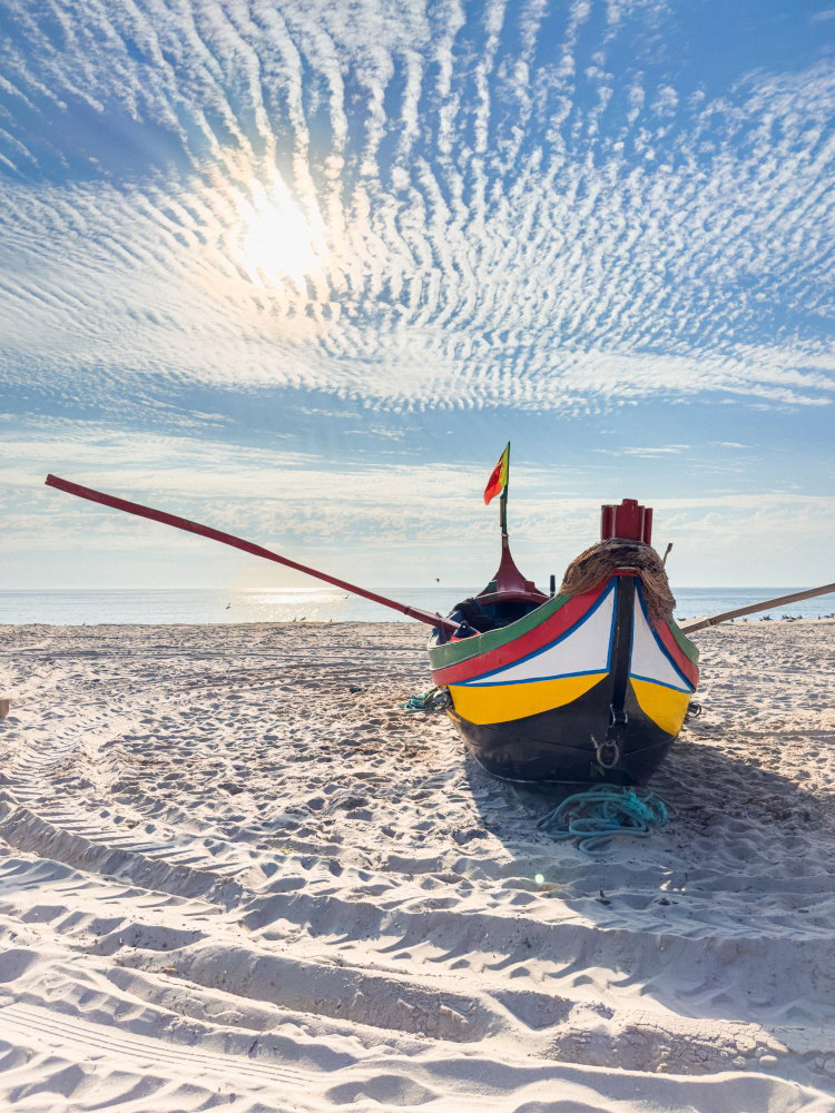 Traditional colorful Portuguese fishing boat used for arte xavega resting on a sandy beach under a dramatic cloudy sky and bright sun during a beautif Traditional colorful Portuguese fishing boat used for arte xavega resting on a sandy beach under a dramatic cloudy sky and bright sun during a beautif